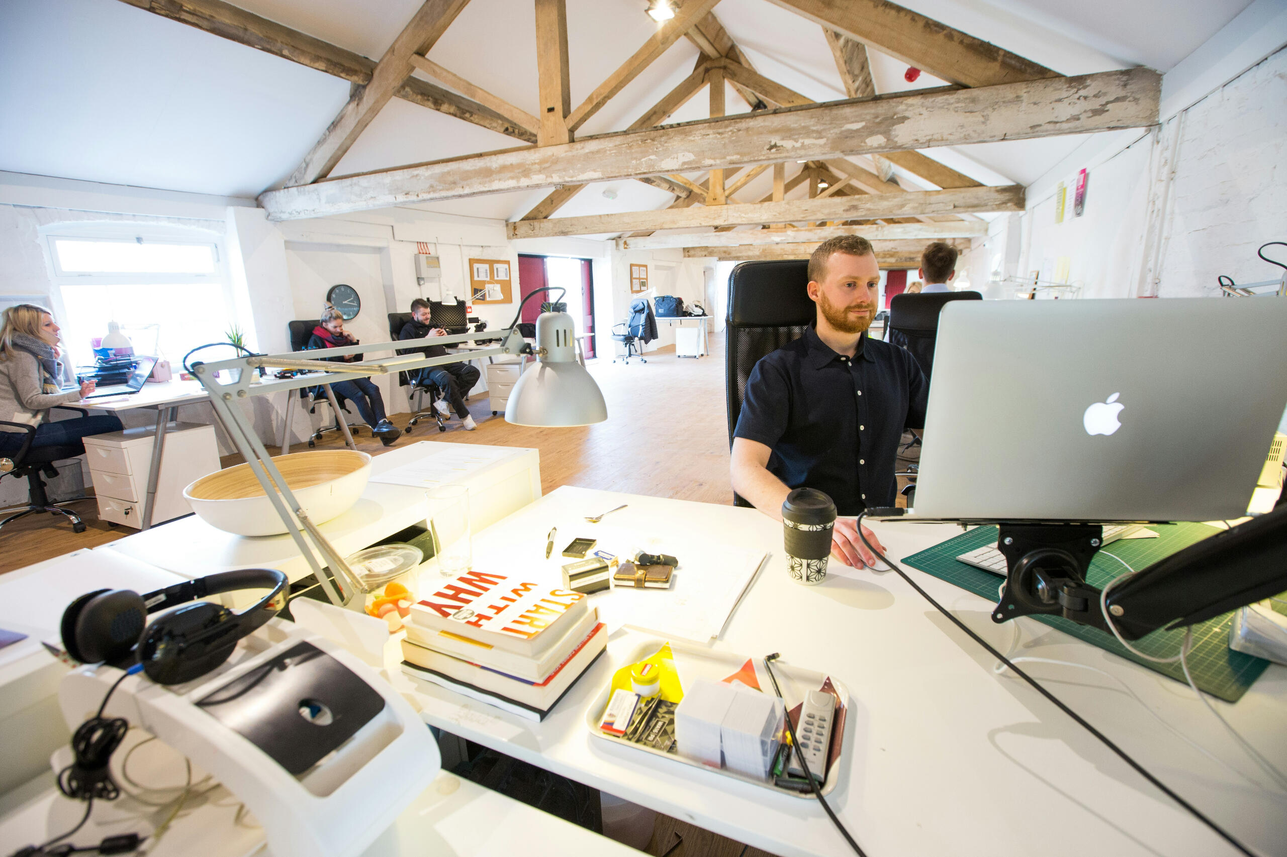 Man sitting at desk in an office looking at a computer A man in a trendy, modern office with other employees, looking at his computer